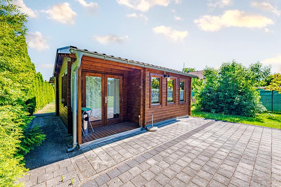 Holzgartenhaus mit Fensterfront auf gepflastertem Platz am Rand des Gartens