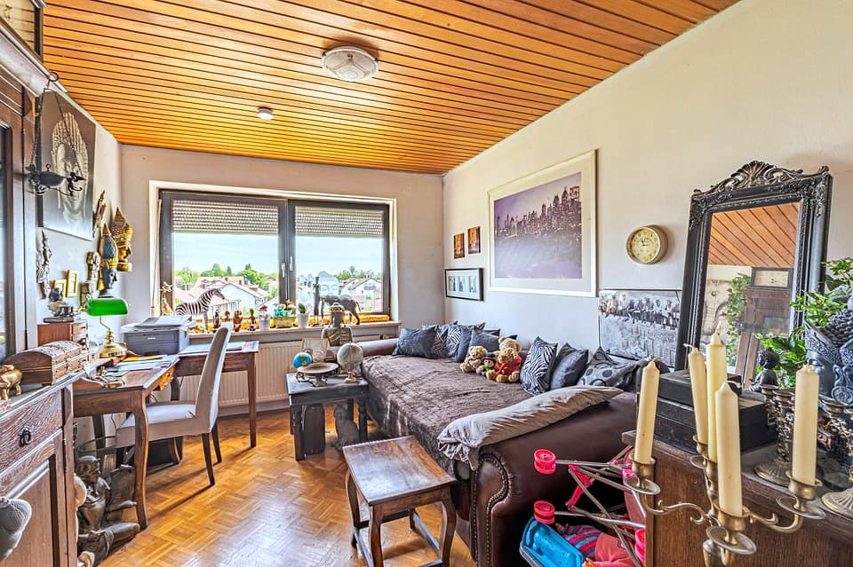 Wohn- und Arbeitszimmer mit Schreibtisch, Sofa und Fenstern ins Grüne