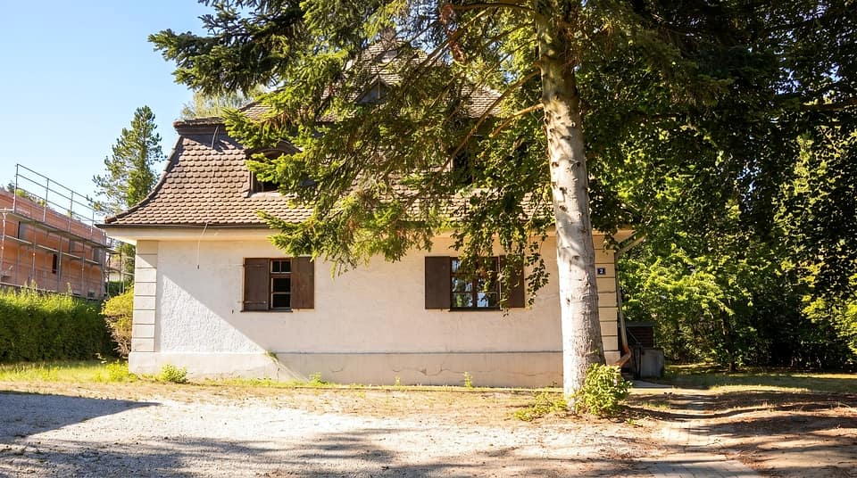 Giebelseite mit Klappläden, davor die gekieste Zufahrt unter alten Bäumen